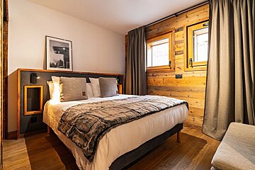 Cozy rustic bedroom featuring a double bed with a textured blanket, wooden walls, grey curtains, and warm lighting.