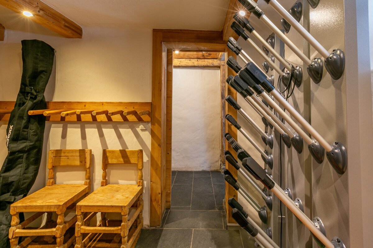 Rustic ski room with a boot drying rack on one wall, wooden benches, a coat rack, and a dark green ski bag, next to a doorway.