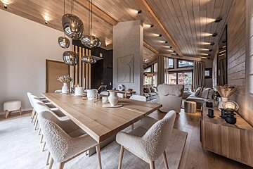 A modern chalet dining and living room with wooden vaulted ceilings, large table, textured chairs, stylish pendant lights, and cozy seating.