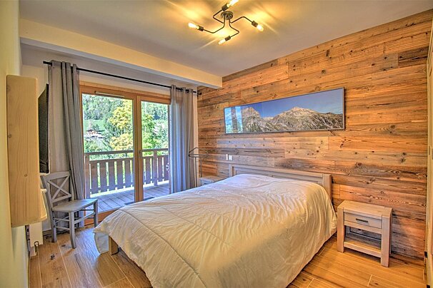 A cozy, rustic bedroom with wooden walls, a white bed, and a balcony showing a lush mountain view under a modern ceiling light.