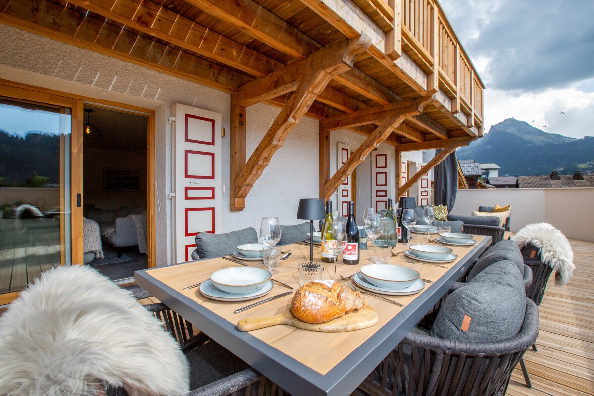 An inviting outdoor terrace with a set dining table, fresh bread, and wine, overlooking picturesque mountains under a timber-framed balcony.