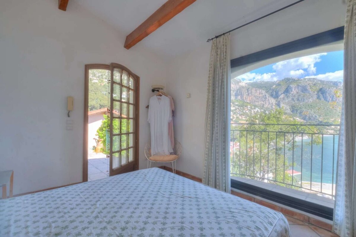Bedroom with a large window overlooking a bay, mountains, and blue sky. A French door leads outside, and wooden beams adorn the ceiling.