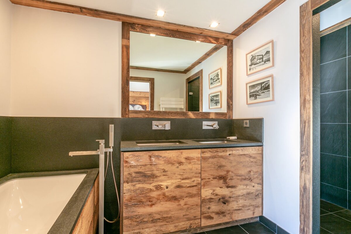 Modern bathroom with rustic wood vanity & mirror, double sinks, stone counter & tub surround. White walls contrasted with dark tiles & wood accents.