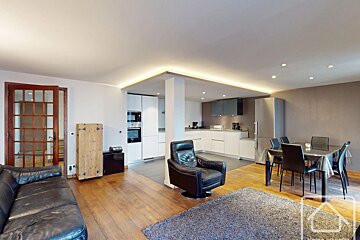A modern open-plan living area with a sleek white kitchen, dining table, black sofa, armchair, and warm wooden floors, featuring recessed lighting.