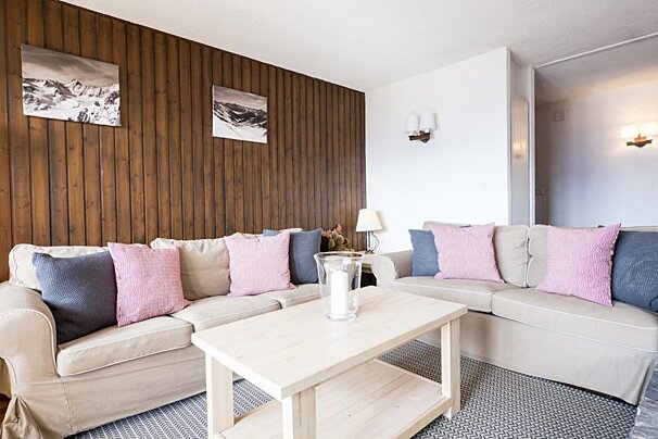 Cozy living room with two beige sofas, pink and blue cushions, a light wooden coffee table, and a wood-paneled wall with mountain art.