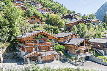 A large wooden house sits on a hillside with other houses