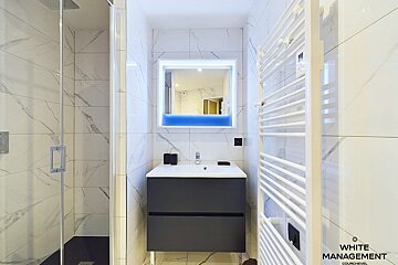 A modern white and gray bathroom featuring a glass shower, dark vanity, backlit mirror with a blue accent, and a white towel warmer.