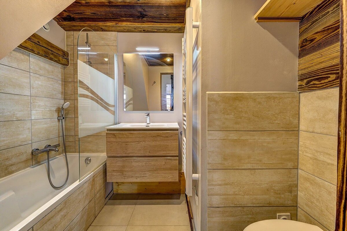 A cozy, modern bathroom featuring a tub/shower, wood vanity, mirror, tiled walls, and rustic wooden ceiling beams.