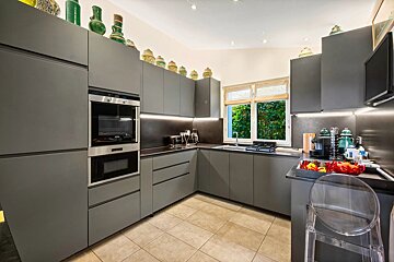 A sleek, modern gray kitchen features built-in appliances, under-cabinet lighting, a window with lush greenery, and decorative pottery atop cabinets.