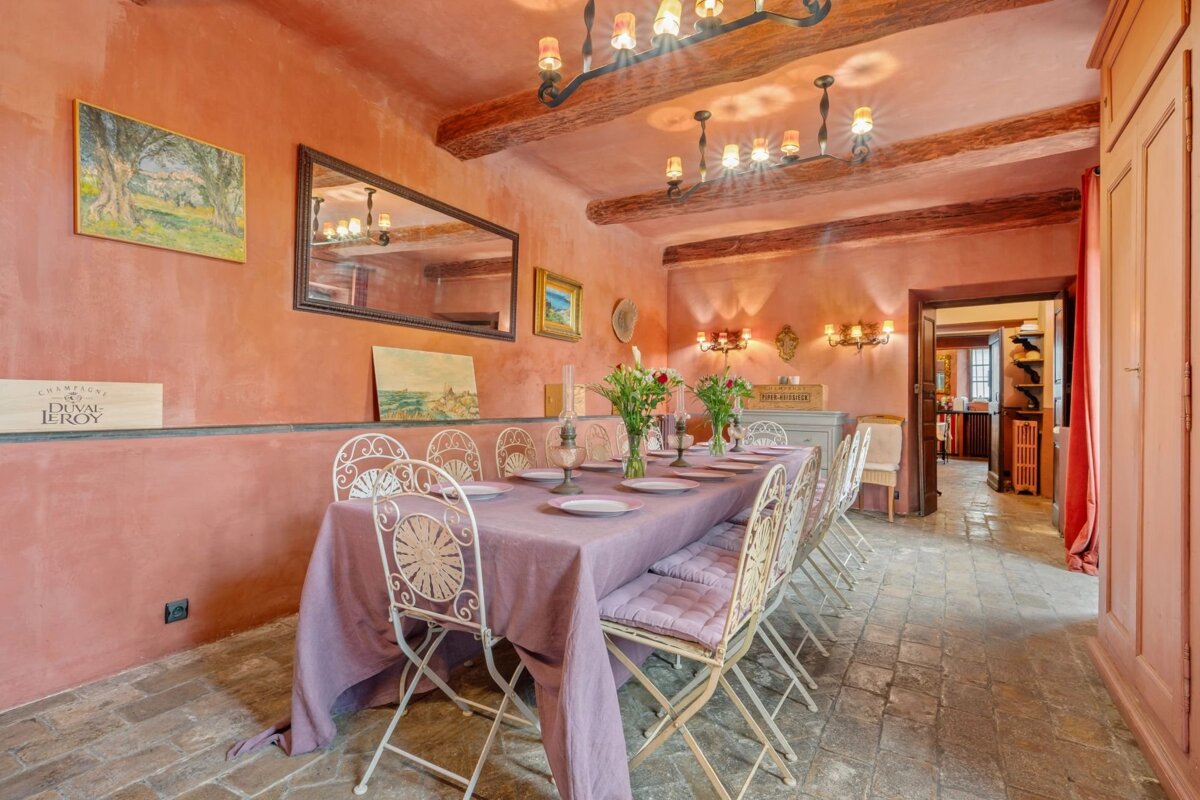 Rustic dining room with terracotta walls, wooden ceiling beams, and a long table set with a purple cloth and white iron chairs.