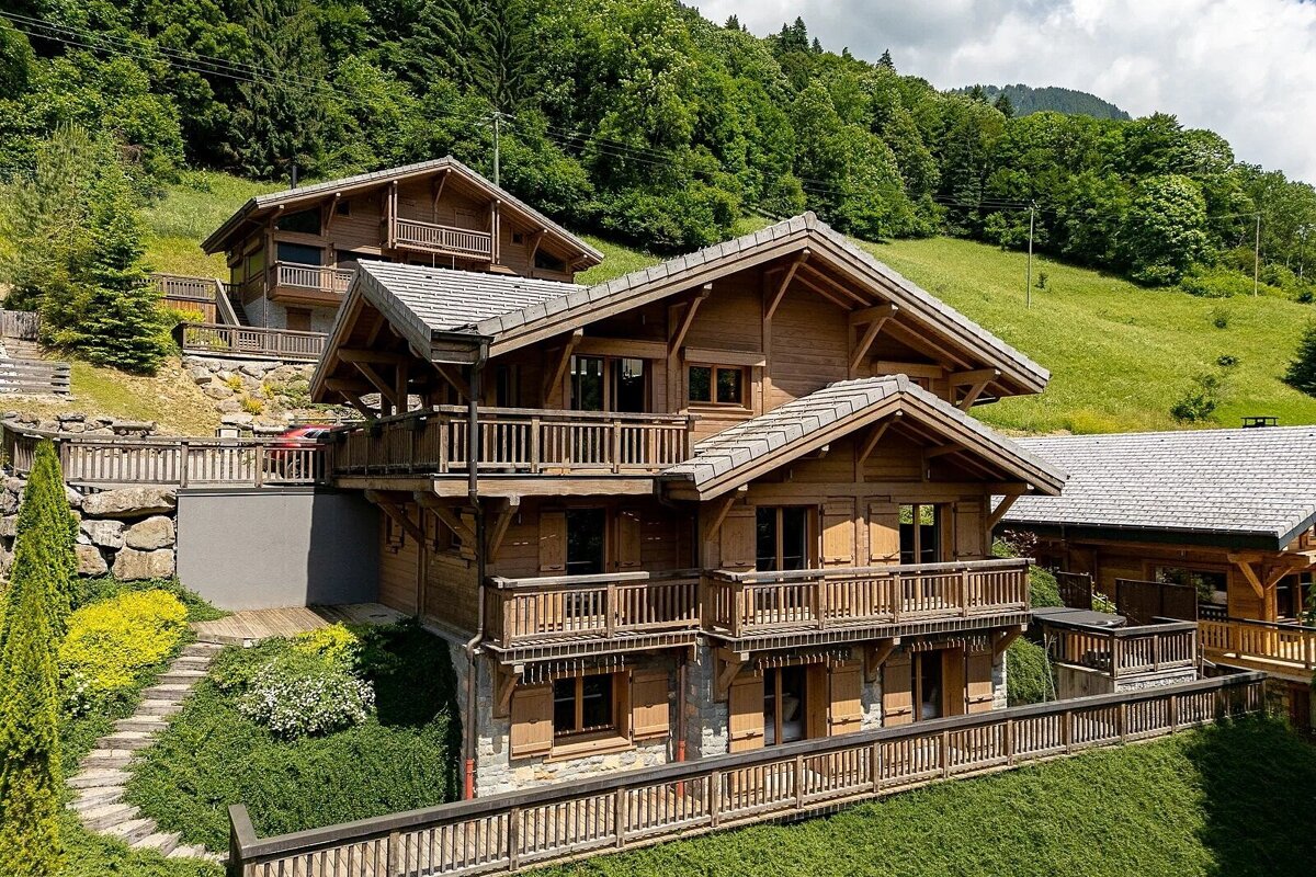 Several wooden chalet-style houses with balconies are nestled on a green, tree-covered mountainside under a cloudy sky.