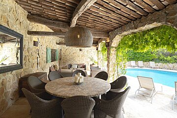 A rustic covered patio with a stone dining table and wicker chairs, opening up to a sunny backyard swimming pool.