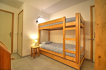A bedroom with a wooden bunk bed with blue/white patterned bedding, a bedside lamp on a wooden table, white walls, and tiled floor.