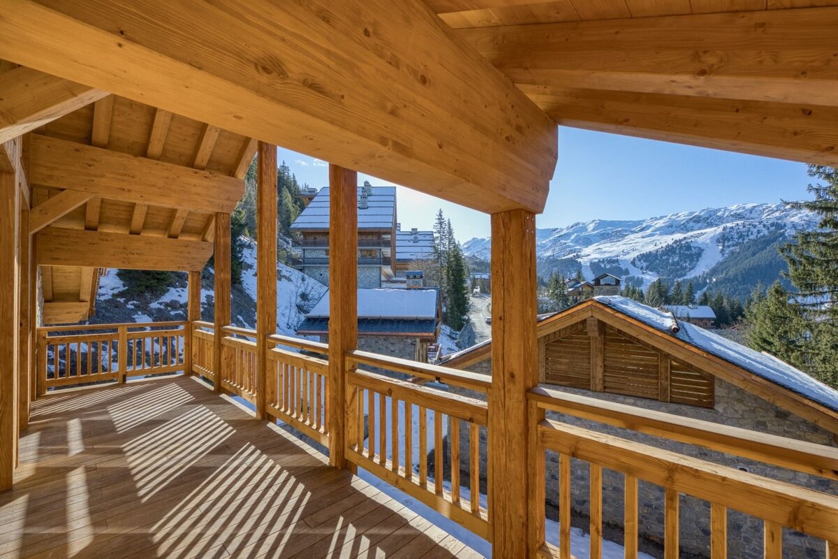 Wooden balcony with sun stripes overlooking a snowy mountain village, chalets, and distant ski slopes under a clear blue sky.