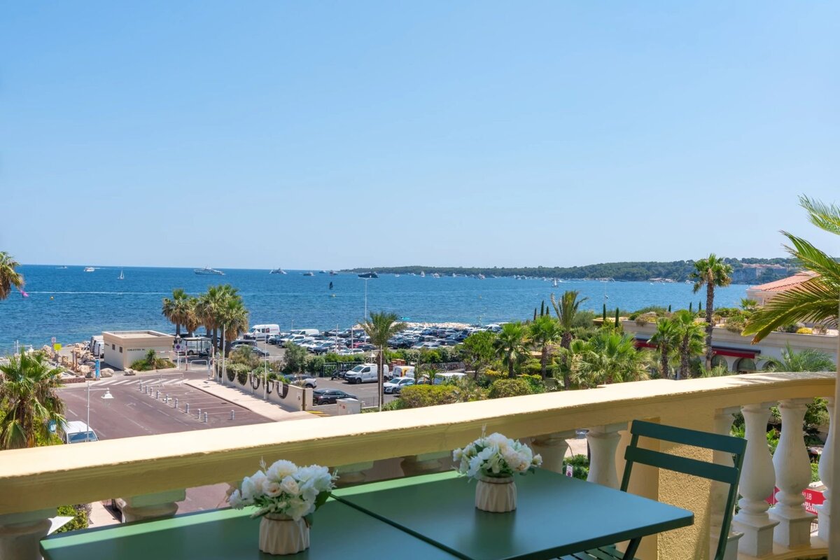 A balcony with a green table and flowers looks out over a sunny blue sea filled with boats and palm trees.