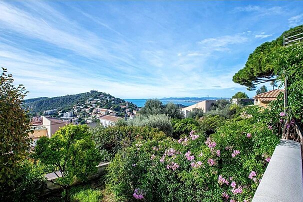 An elevated view of a charming coastal town nestled on hills, overlooking the blue sea. Lush green gardens with pink flowers frame the foreground under a vast sky.