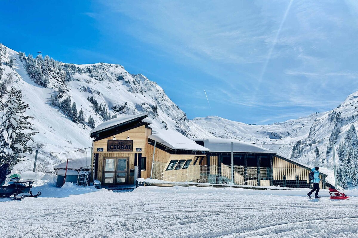 A large wooden chalet with balconies and stone foundation, covered in snow, sits in a sunny mountain village under a clear blue sky.