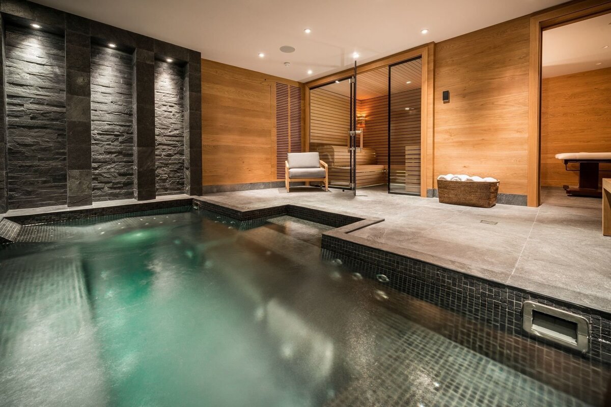 A modern, luxurious spa area featuring a dark-tiled hot tub, a glass-fronted sauna, and rich wood paneling with a stone accent wall.