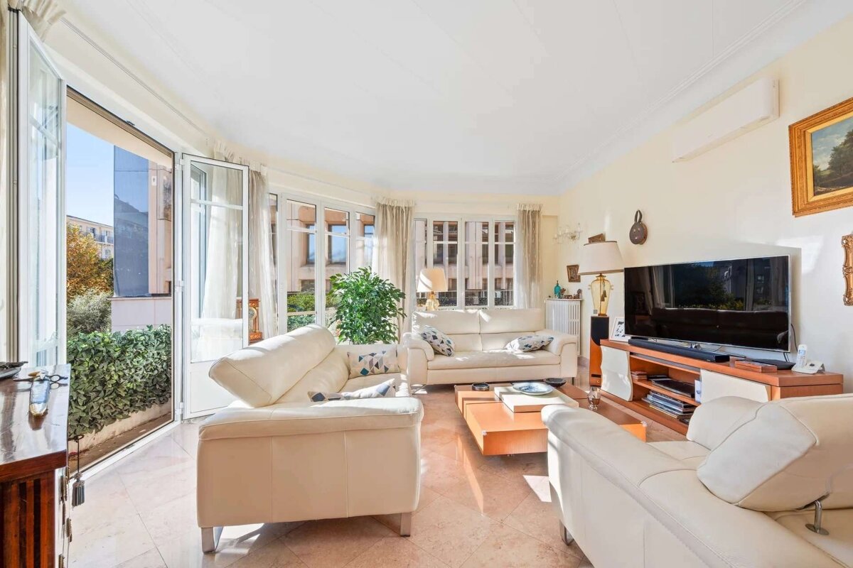 Bright living room with cream sofas, a TV, and large windows opening to a green outdoor space.