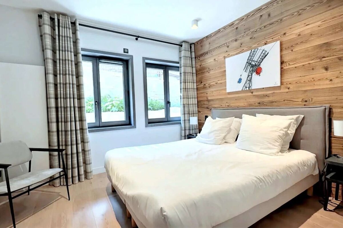 A cozy, modern bedroom with a large white bed, wood accent wall, ski lift art, and windows with plaid curtains.