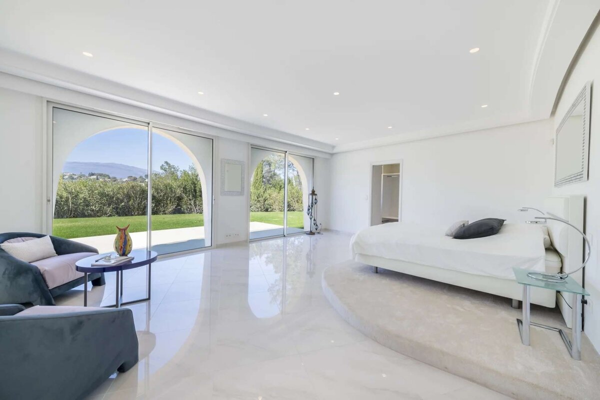 Bright, modern bedroom with large arched windows to a lush garden. White bed, armchairs, and glossy floors create a serene, spacious feel.