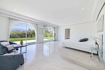 Bright, modern bedroom with large arched windows to a lush garden. White bed, armchairs, and glossy floors create a serene, spacious feel.