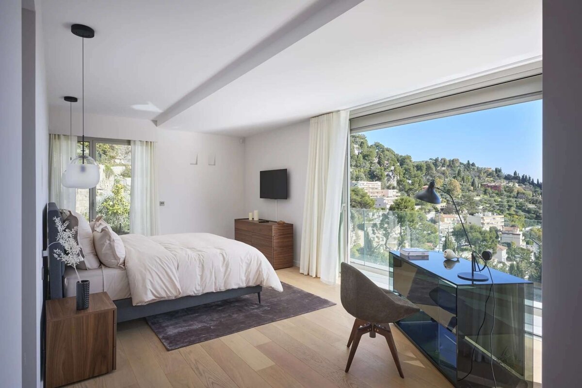 A modern bedroom with a bed, desk, and a large window overlooking a scenic hillside with trees and buildings under a clear sky.