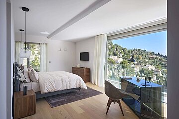 A modern bedroom with a bed, desk, and a large window overlooking a scenic hillside with trees and buildings under a clear sky.