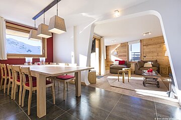 Bright chalet interior with a wooden dining table and red-cushioned chairs, opening into a cozy wood-paneled living area.