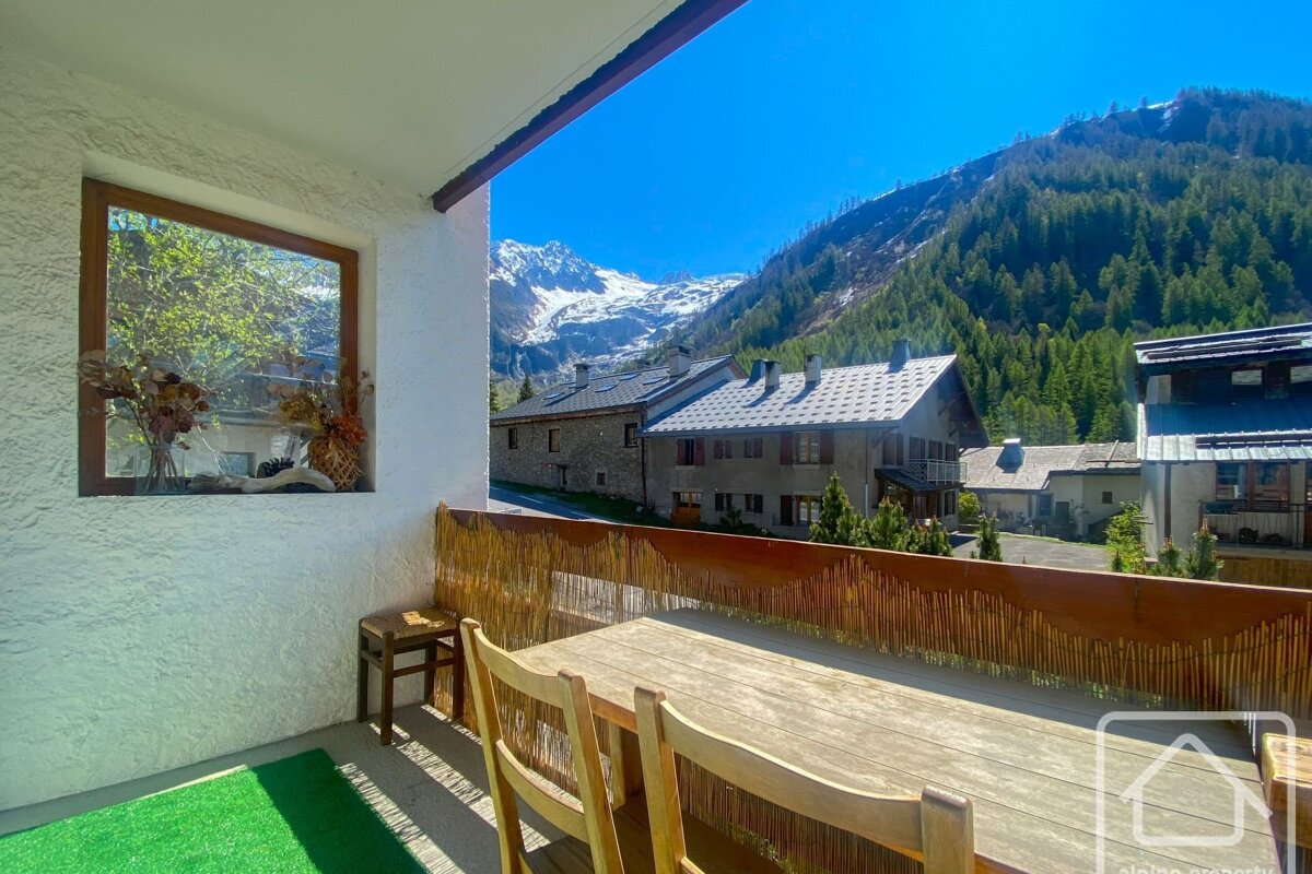 Sunny balcony with wooden table and window looking out to an alpine village, forested hills, and distant snow-capped mountains under a blue sky.