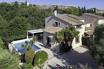 An aerial view of a beige villa with a swimming pool, patio, and lush trees, with larger buildings in the background.
