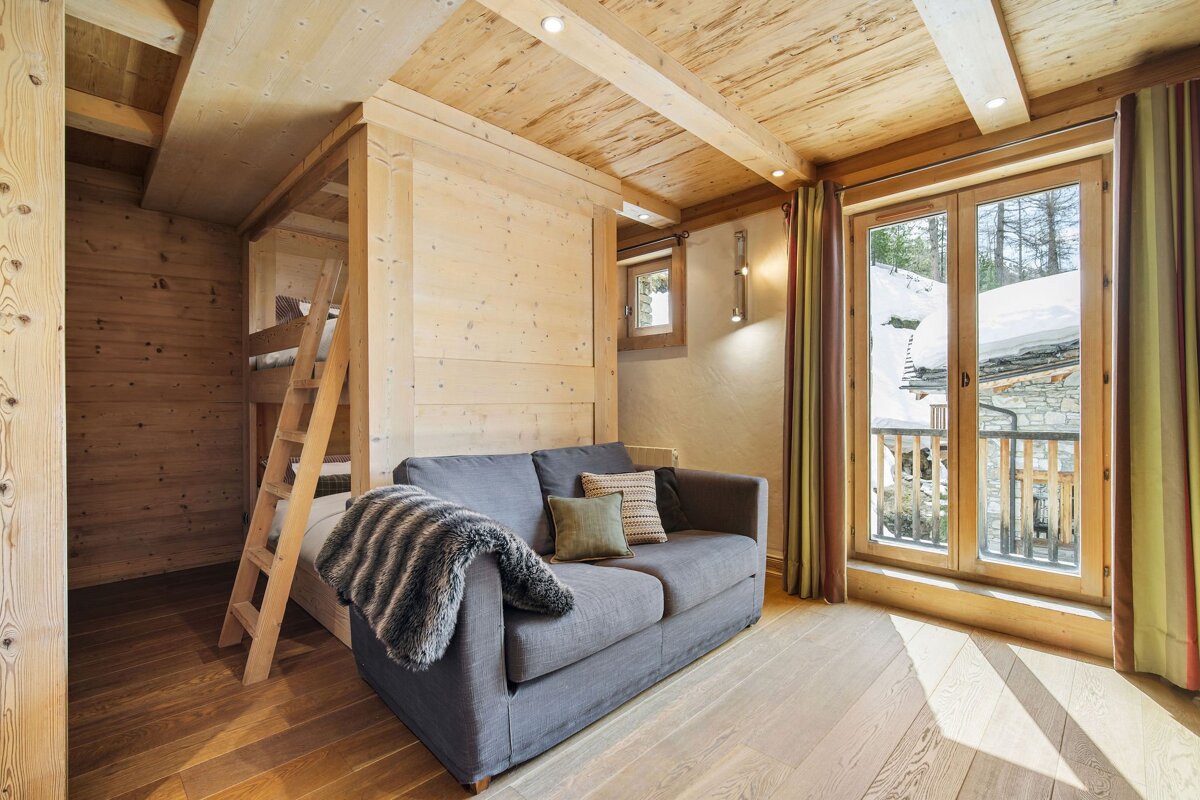Cozy wooden chalet room featuring a grey sofa, wooden bunk beds, and glass doors looking out onto a snowy winter landscape.