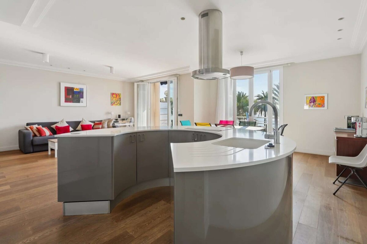 A modern open-plan living space featuring a curved kitchen island, a cozy living room with a sofa, and a balcony overlooking palm trees and the sea.