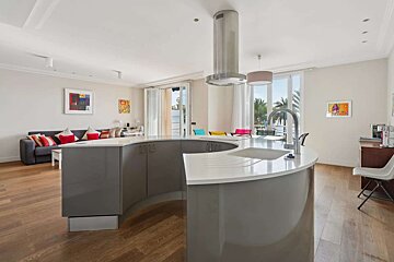 A modern open-plan living space featuring a curved kitchen island, a cozy living room with a sofa, and a balcony overlooking palm trees and the sea.