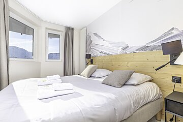 A cozy bedroom with a large bed, wooden headboard, and mountain mural. Scenic snowy mountain views from two windows.