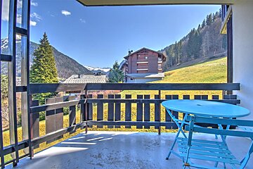 A sunny balcony with blue furniture offers a scenic view of green mountain slopes, distant houses, and snowy peaks under a clear blue sky.