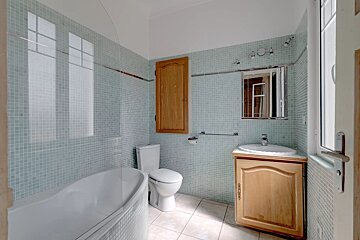 A bathroom with light blue mosaic tiles, a white bathtub, toilet, and wooden vanity with a sink. A wooden wall cabinet and window are also visible.