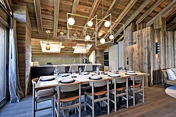 A chic, spacious dining room and open kitchen in a chalet style, featuring a long table, rustic wooden interior, and modern geometric chandeliers.