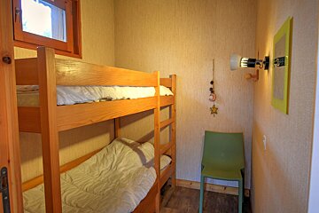 A small, cozy bedroom features wooden bunk beds, a window, a green chair, and celestial wall decor, illuminated by a wall lamp.