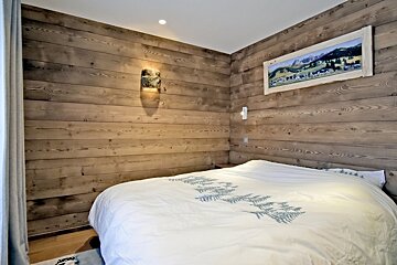 Cozy cabin bedroom with rustic wood-paneled walls, a bed with a pine-tree patterned duvet, and a scenic framed painting on the wall.