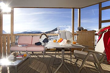 A sunny balcony deck with outdoor furniture, winter clothing, fresh fruit, and drinks, offering a panoramic view of snow-capped mountains.