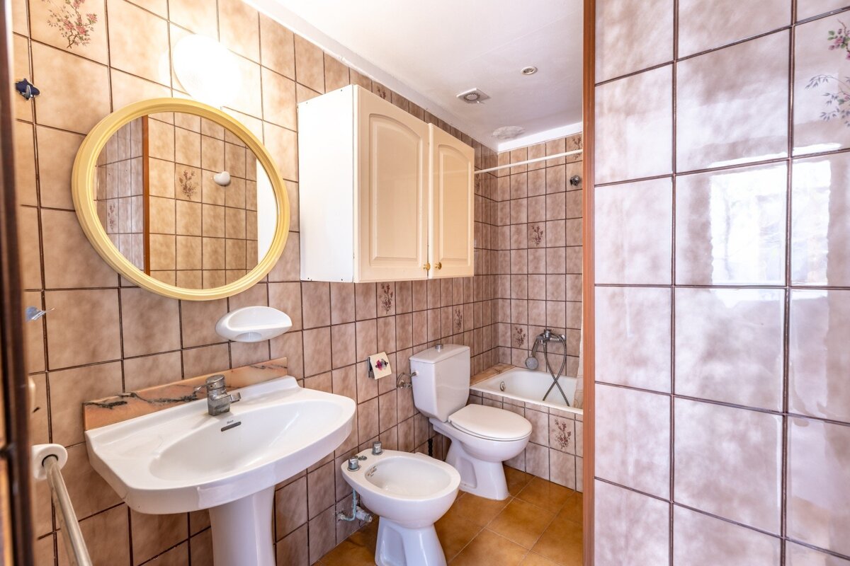 A beige-brown tiled bathroom with a pedestal sink, round mirror, toilet, bidet, and a bathtub with shower. White cabinet above.