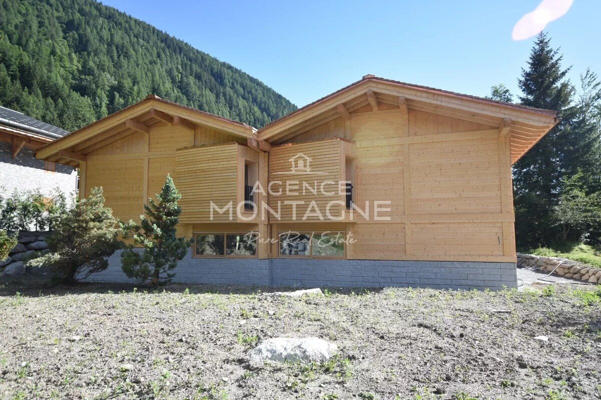 Modern wooden chalet with a stone foundation, set against a lush, forested mountain under a clear blue sky.