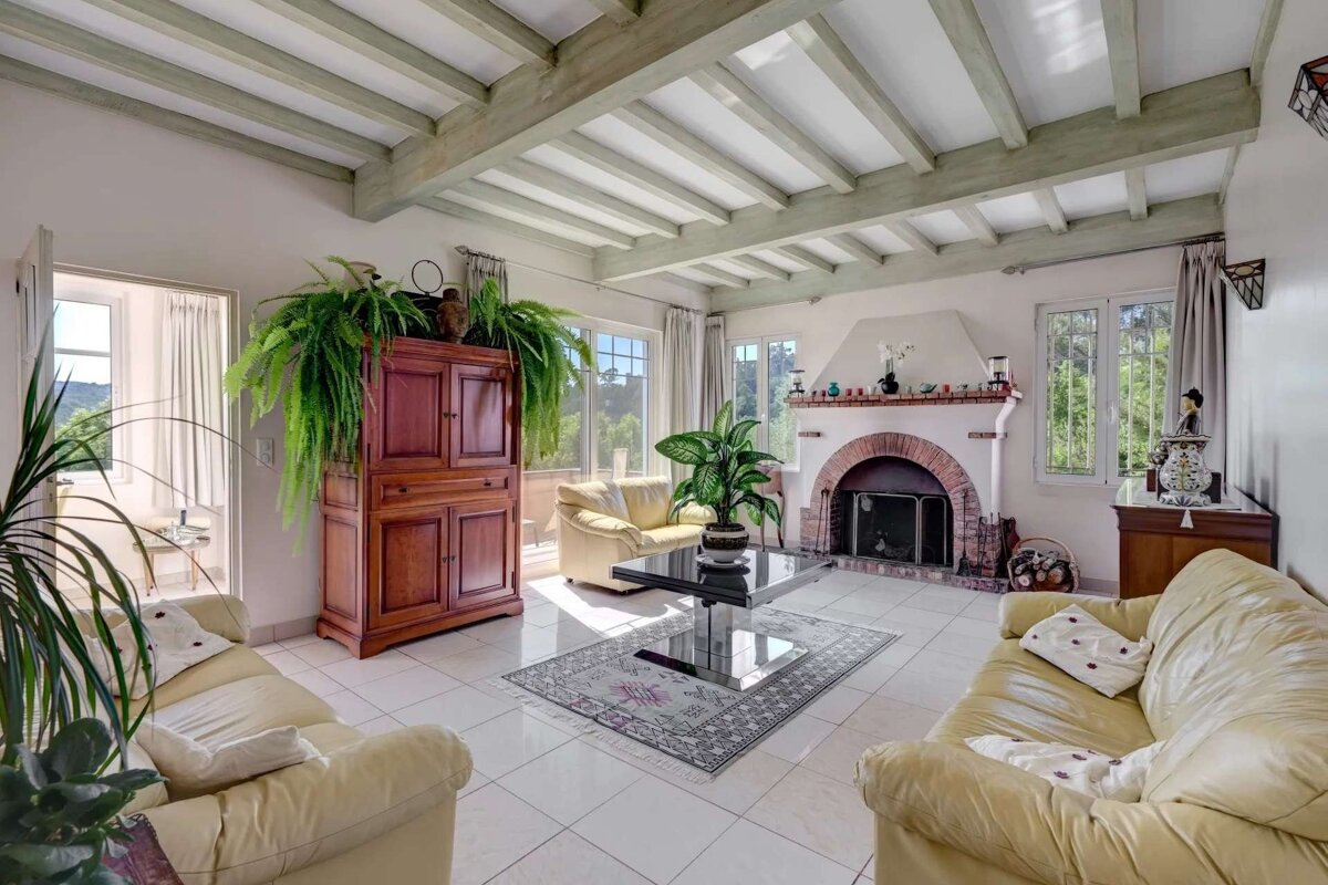 A bright living room with cream sofas, white tiled floor, exposed beam ceiling, brick fireplace, and large windows looking out to lush greenery.