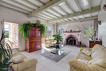 A bright living room with cream sofas, white tiled floor, exposed beam ceiling, brick fireplace, and large windows looking out to lush greenery.