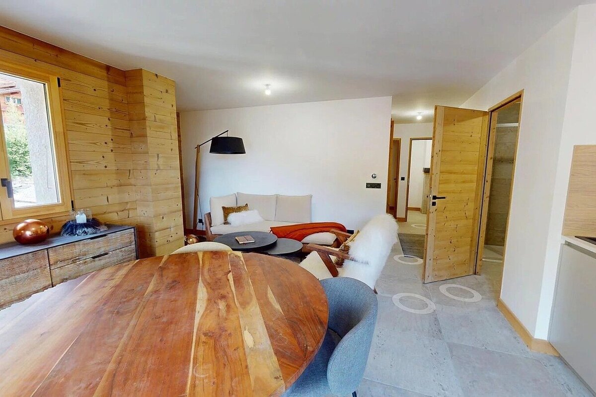 A modern living and dining area features wood paneling, a round wooden table, a light sofa with an orange throw, and a stylish floor lamp.