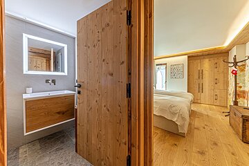 Modern bathroom with vanity & lit mirror, separated by a folding wooden door from a cozy bedroom with twin beds, wood floors, & wardrobes.