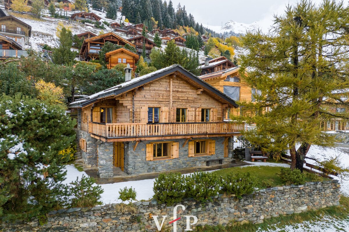 A beautiful wooden and stone chalet nestled on a snowy mountain slope surrounded by alpine cabins and pine trees.