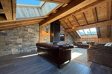A sunlit, rustic chalet living room with stone walls, exposed wooden beams, multiple skylights, and brown leather sofas centered around a modern fireplace.
