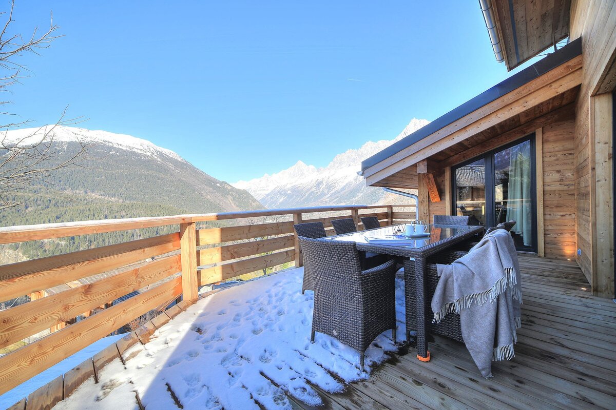 A wooden deck with outdoor furniture and patches of snow, offering a panoramic view of snow-capped mountains and a valley under a clear blue sky.